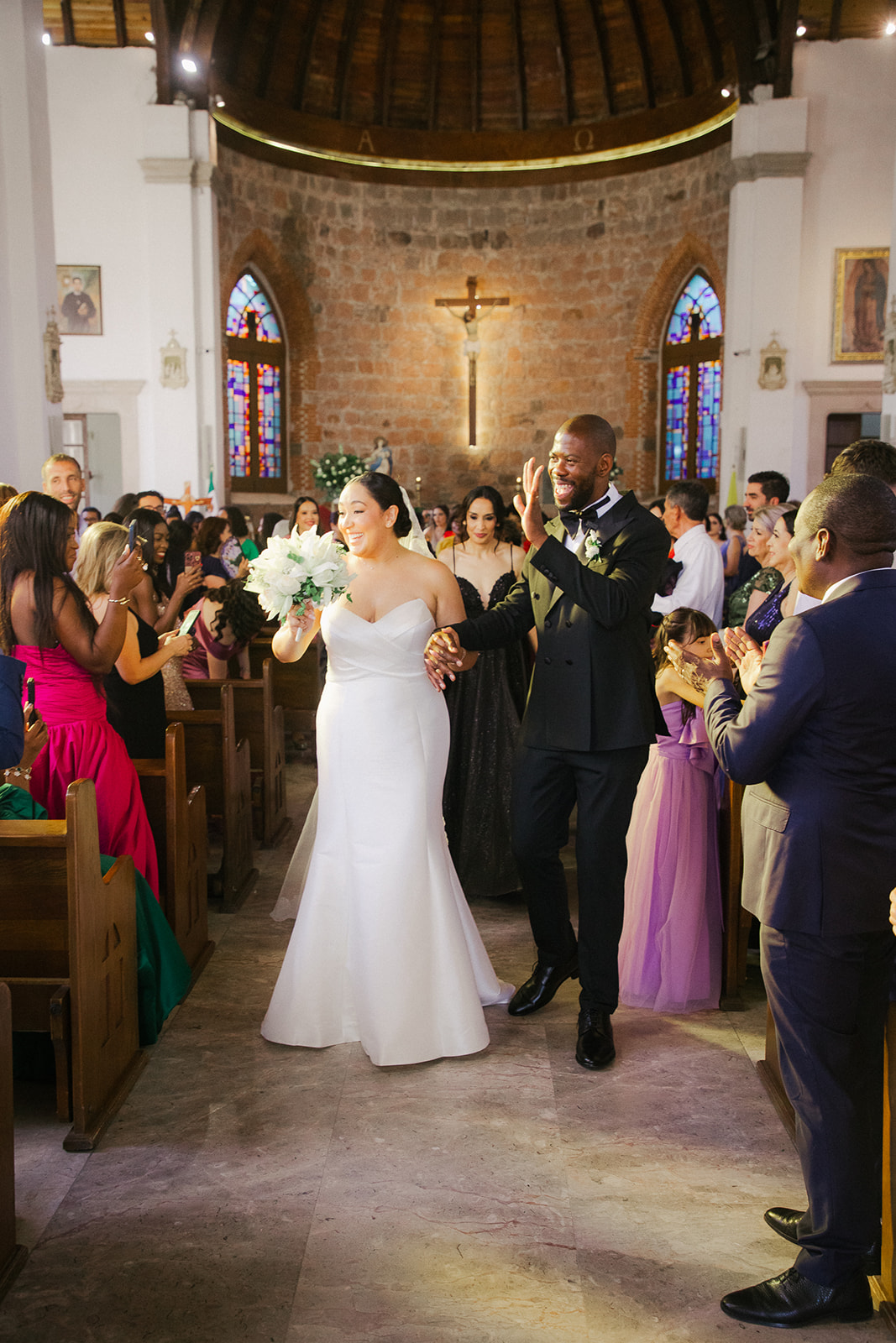 Bride and groom walk hand in hand down the church aisle after their ceremony as guests applaud — the bride smiling with a white bouquet, the groom in a black tuxedo greeting the crowd inside a colonial stone church; documentary wedding photography by David Tovar in Mexico